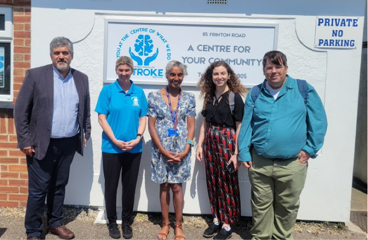 Five people standing, posing for a photo in front of the stroke community centre sign