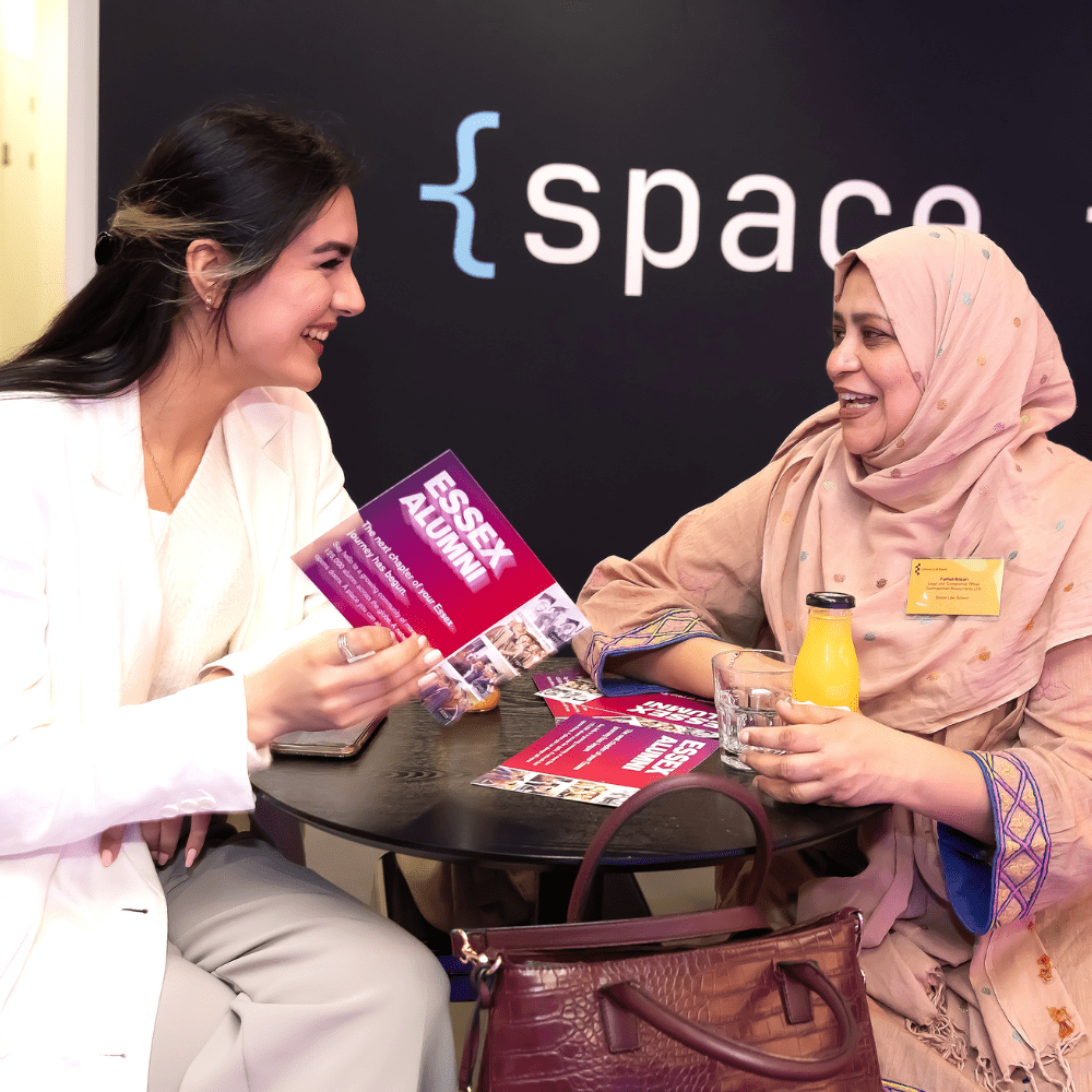 A prospective student wearing a white suit chats with a graduate wearing a pink hijab and outfit, they are sat on high chairs at a posy table. 