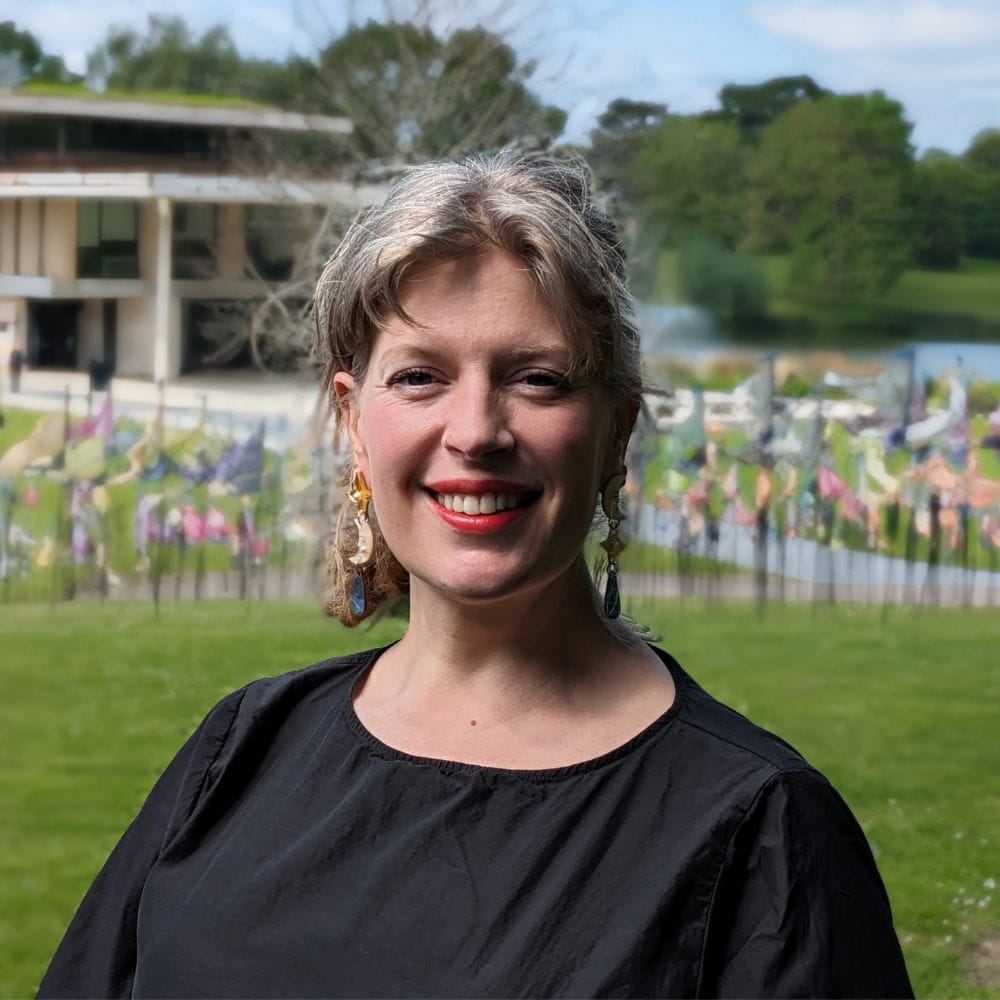 Dr Sarah Perry, smiling and looking relaxed with a colourful backdrop of flags, planted in the ground on a grassy hill