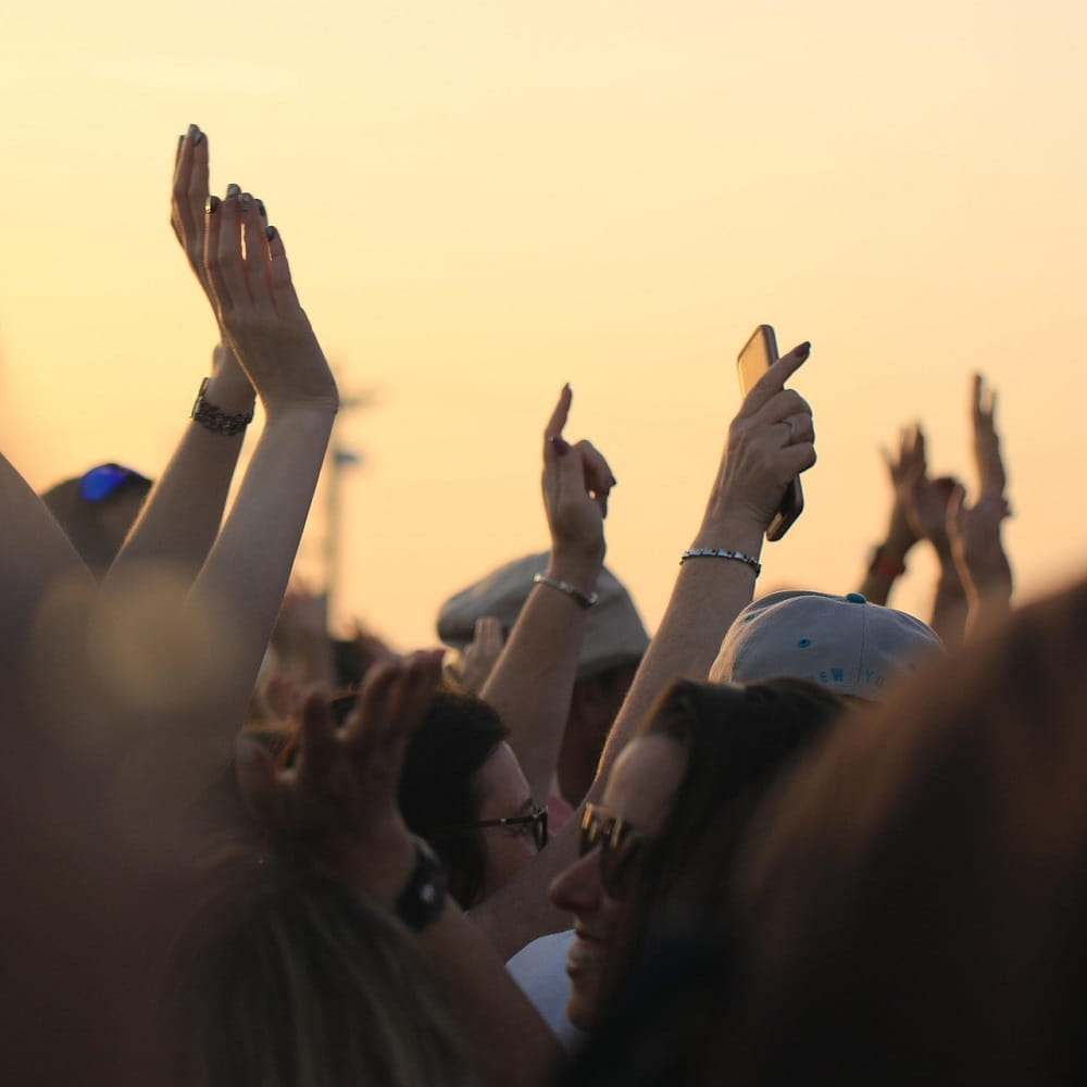 A crowd of people waving their hands in the air, with a sunset glow in the sky in the background.
