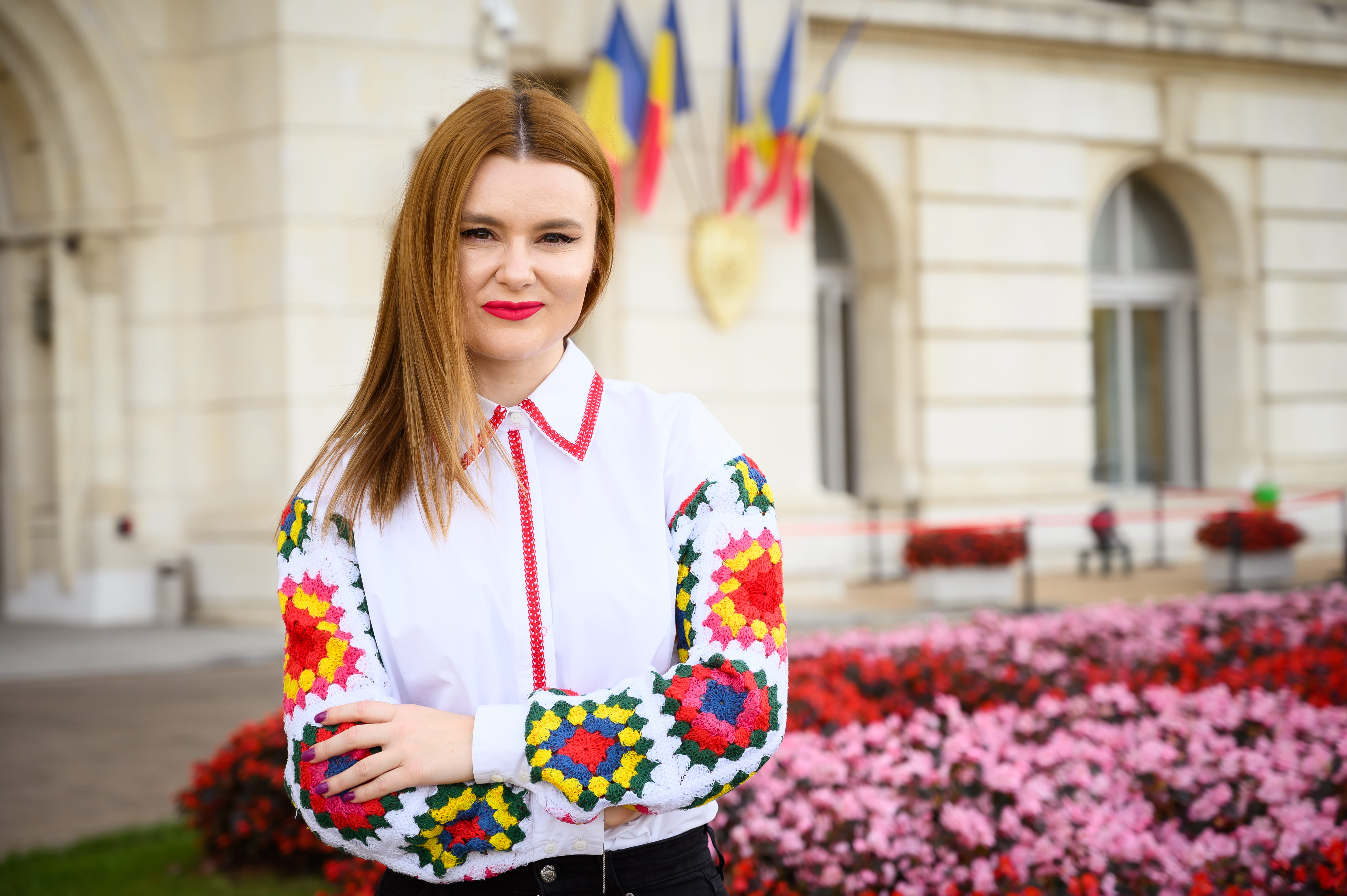 simina wearing a white shirt with colourful sleeves, stood in front of an official building with the Romanian flags flying. 