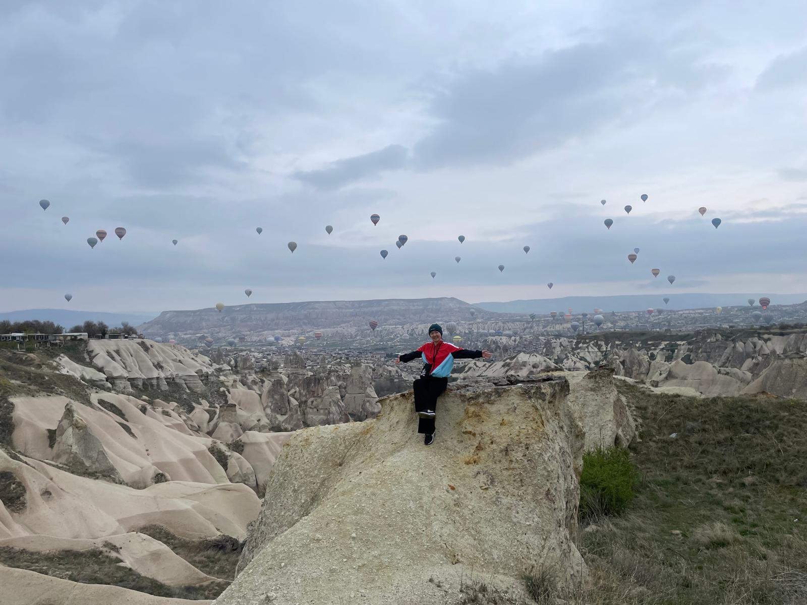 MA Curating 2024 graduate, Hiromi Horiuchi, stands atop a mountain surrounded by hot air balloons