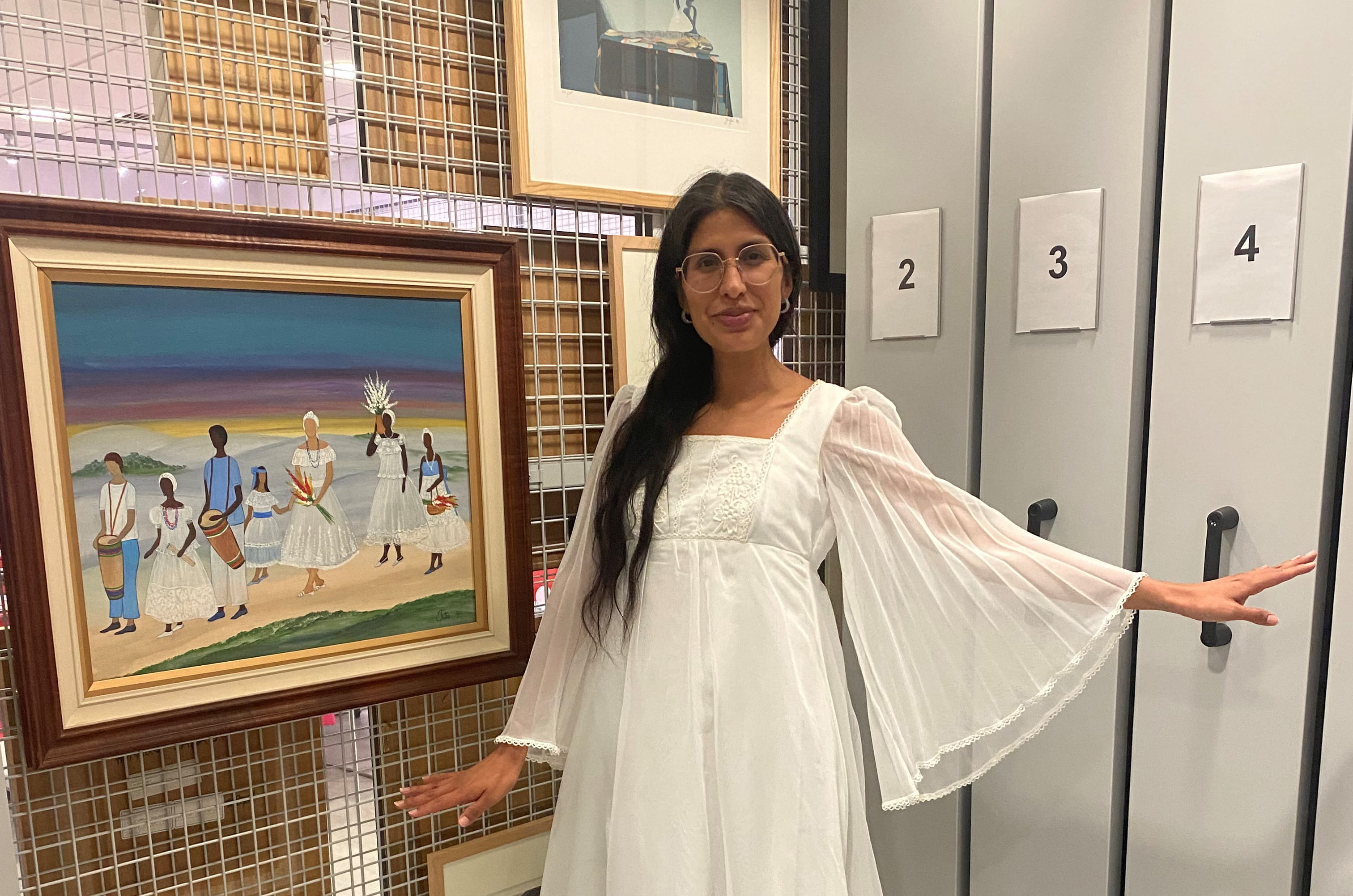 Assistant Curator of ESCALA and the University Art Collection, Gisselle Giron wears a white Easter Dress and stands next to Tiita Machado’s “Á caminho da festa de Iemanjá” (1996) in the ESCALA Teaching and Research Space.