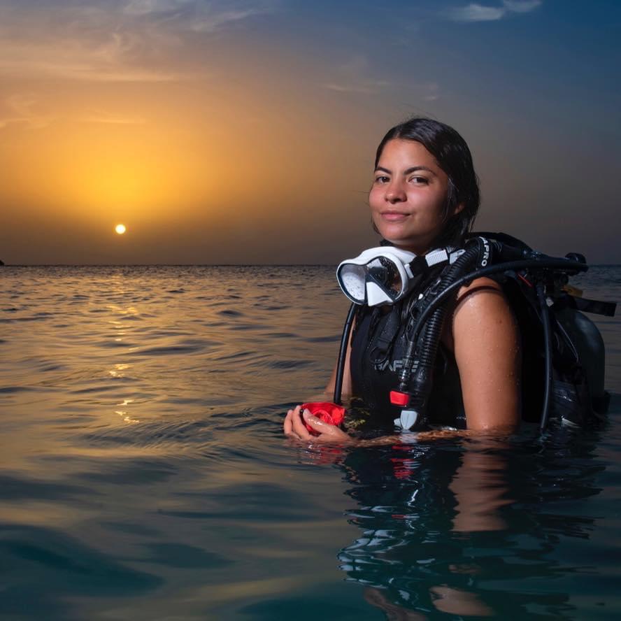 Damaris in the sea wearing scuba diving kit, the sun is setting behind her