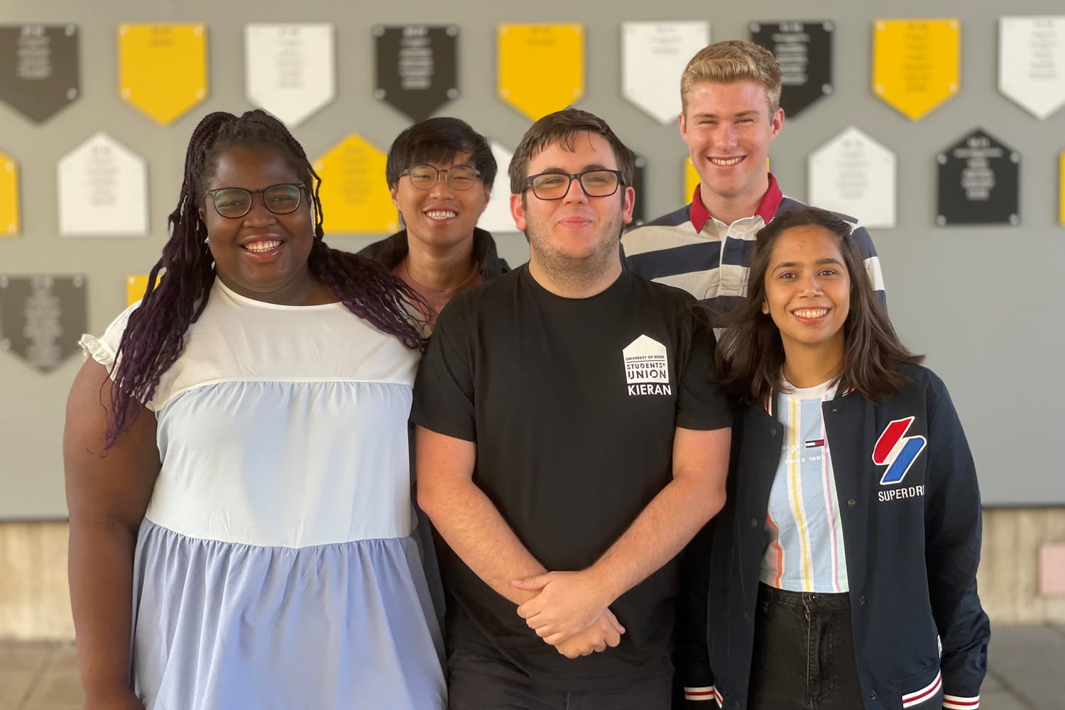 Five Students' Union leaders for 2023-24 smile in front of the Wall of Fame. Each plaque on the wall bears the names of leaders of the SU for an historical year.