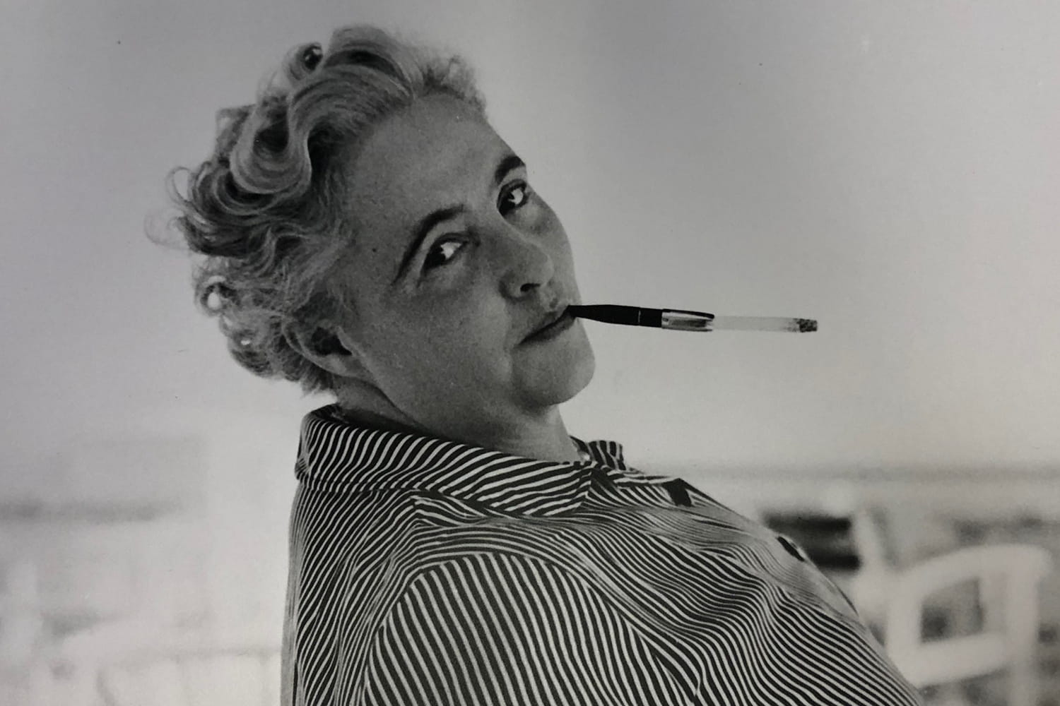 A black and white photo of Margery Allingham looking absolutely fabulous with her coiffed hair and peering over her shoulder while balancing a cigarette holder with lit cigarette between two fingers,