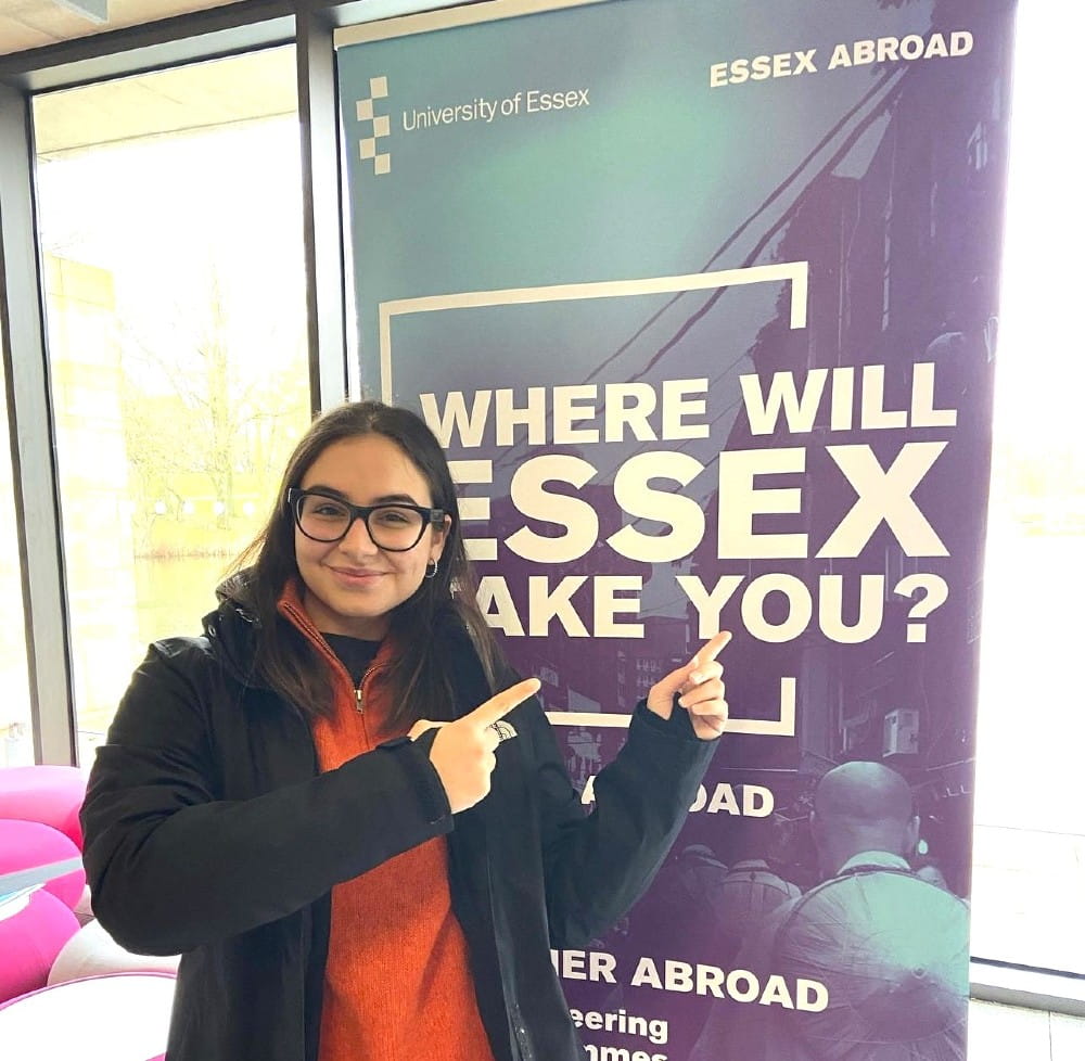 A student ambassador stands in front of a sign which reads "where will Essex take you"