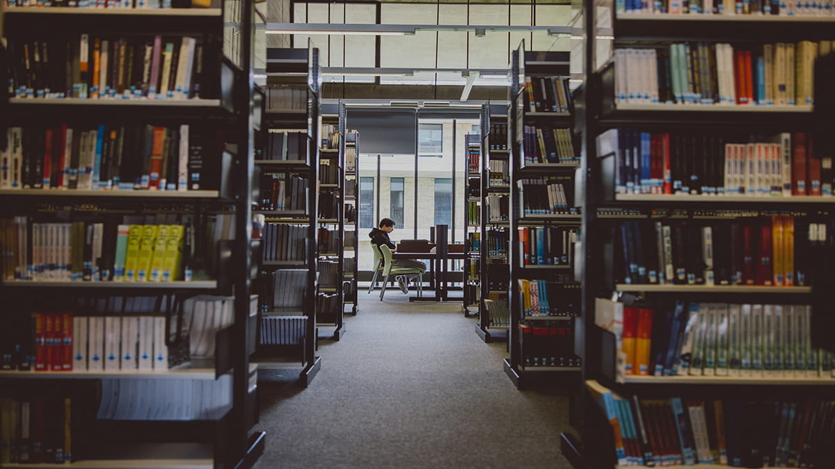 Rows of bookshelves in the Albert Sloman Library. The bookshelves have a centre aisle, and at the end of the aisle is a student sat at a desk studying. 