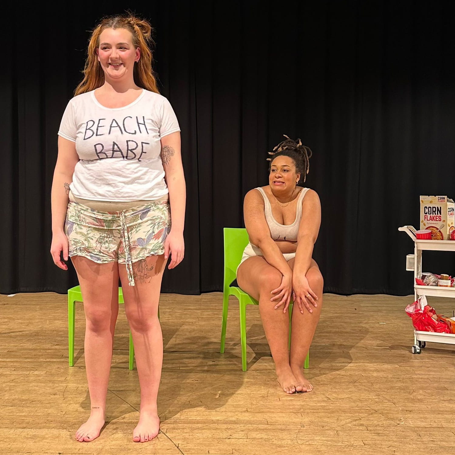 Amy Stephenson-Yankuba and Rio Montana Topley perform Nasty on a theatre stage. Rio is wearing a t shirt with "beach babe" crudely written on it.