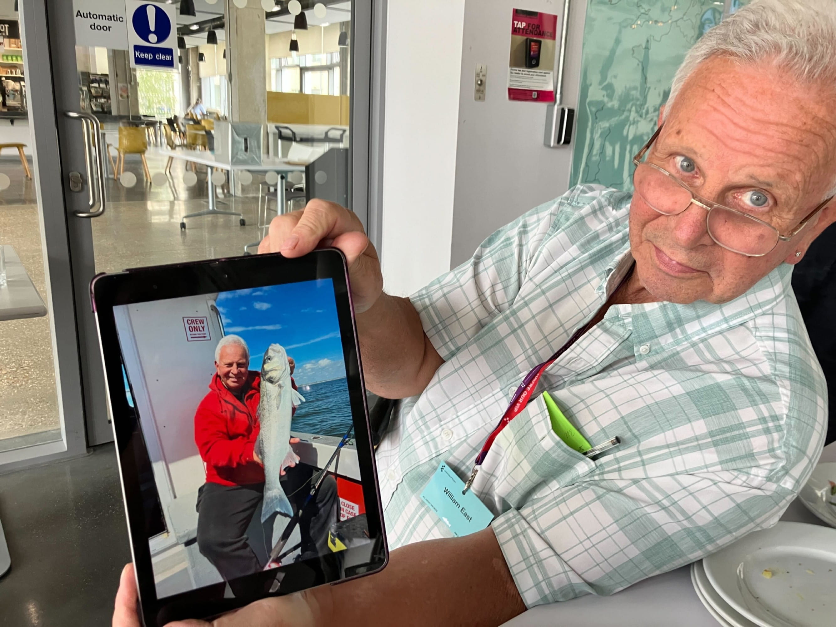 A man with glasses and white hair, leaning towards the camera, holding up a tablet showing a photo of him holding a large fish.