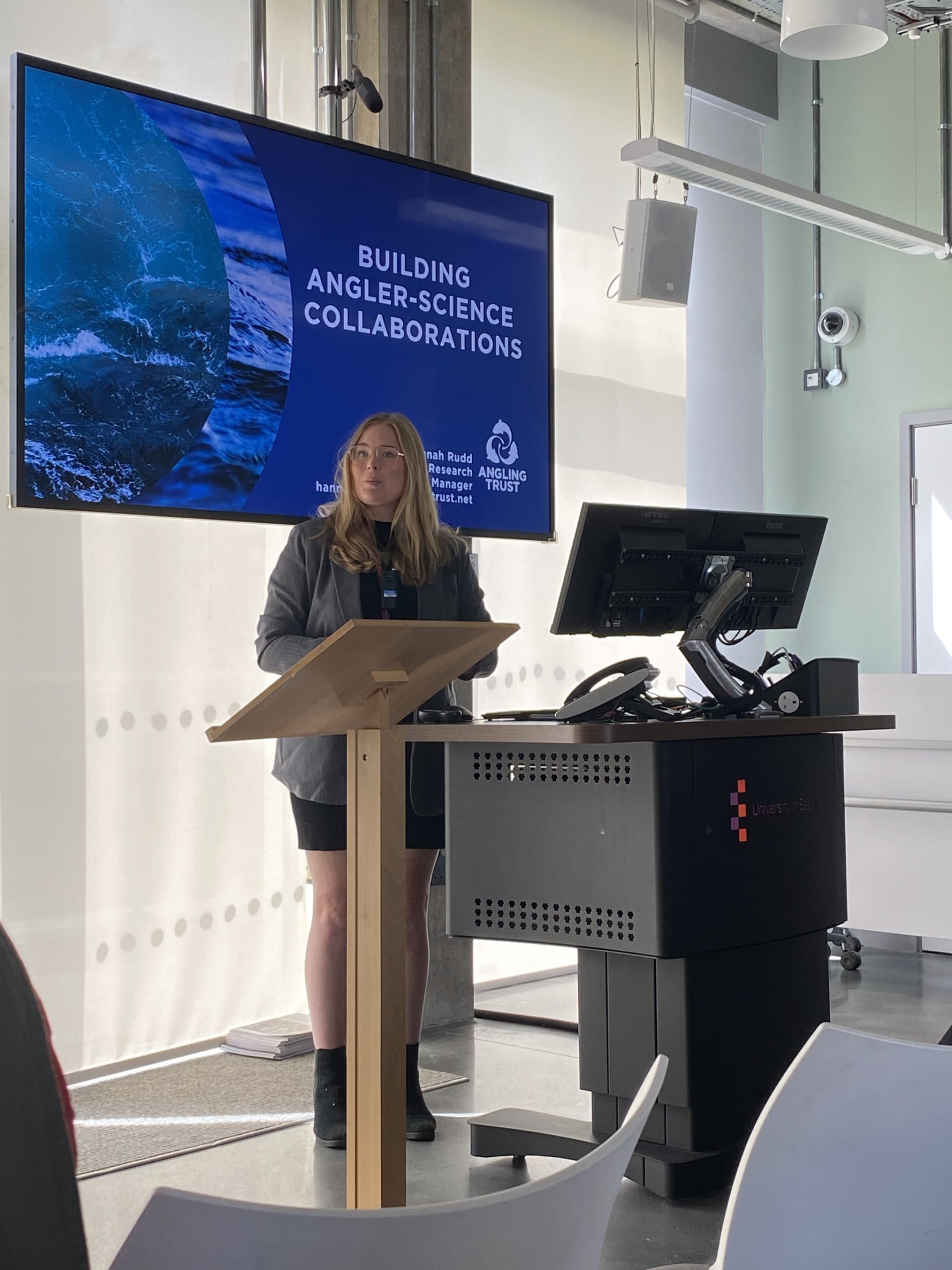 A woman standing in front of a podium and computer screen, talking to the room. Behind her on a large screen is her slide, titled "Building Angler-Science Collaborations."