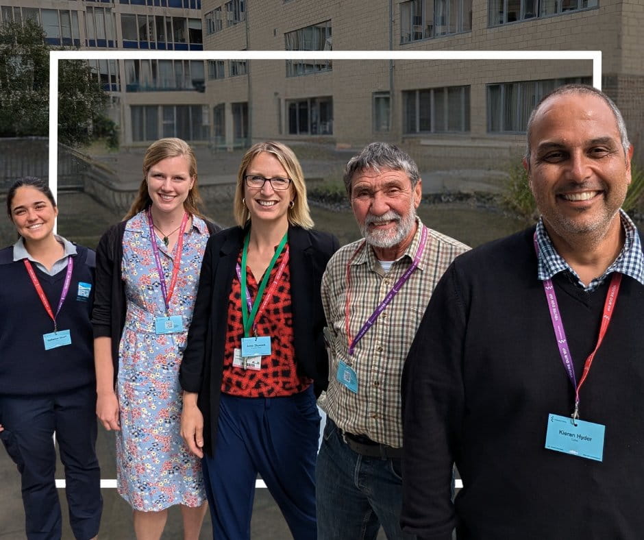 Three women and two men, all with lanyards, standing outside in a row and smiling at the camera.