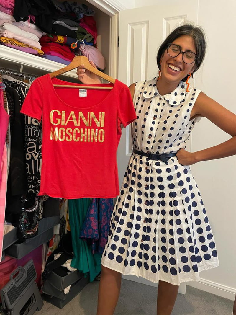 Assistant Curator of ESCALA and the University Art Collection, Gisselle Giron wears a white and black spotted dress and stands holding a red Gianni Moschino tee-shirt on a hanger next to her closet.