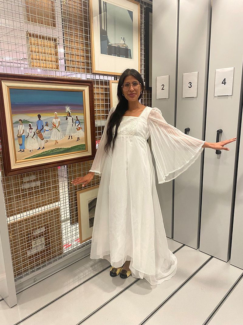 Assistant Curator of ESCALA and the University Art Collection, Gisselle Giron wears a white Easter Dress and stands next to Tiita Machado’s “Á caminho da festa de Iemanjá” (1996) in the ESCALA Teaching and Research Space.