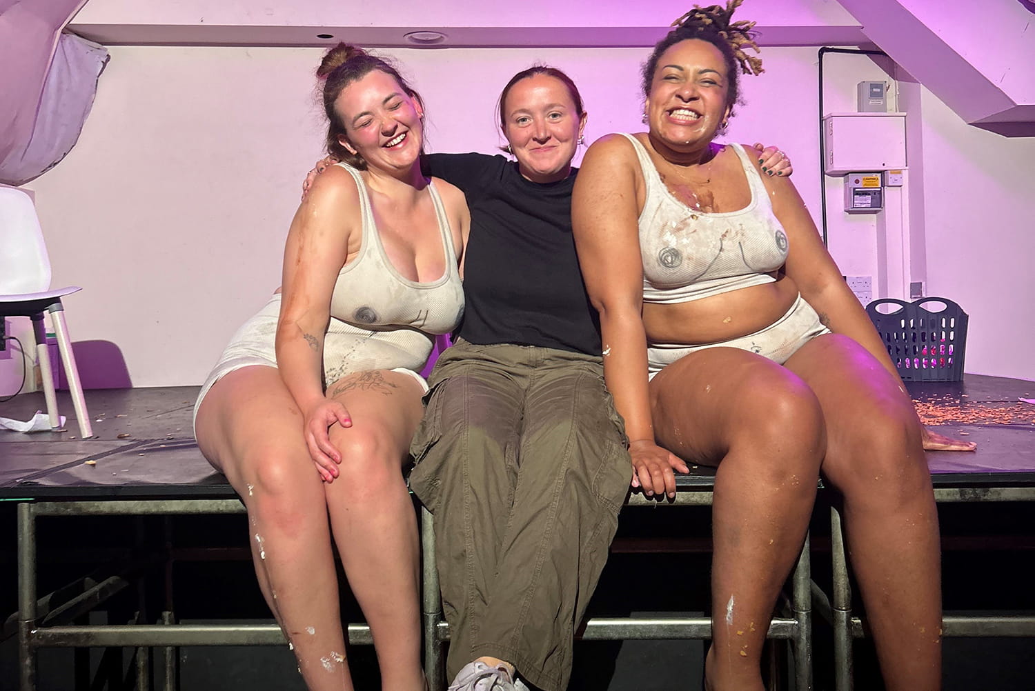 Rio, Amy and Katja of Succulent Theatre sit on a stage hugging together.