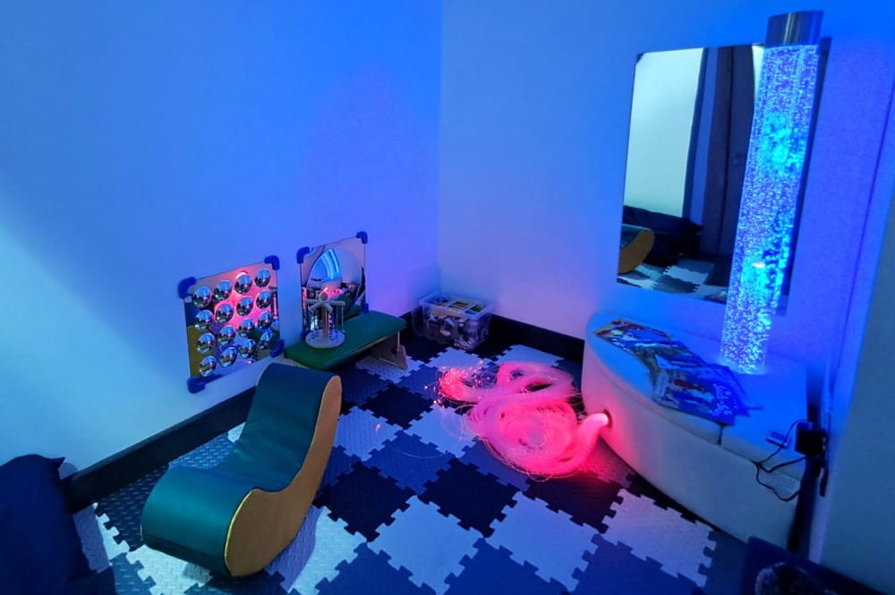 A sensory room with the lights turned down, with square patterned floor, mirror, bubble light tower, and some toys and seating.