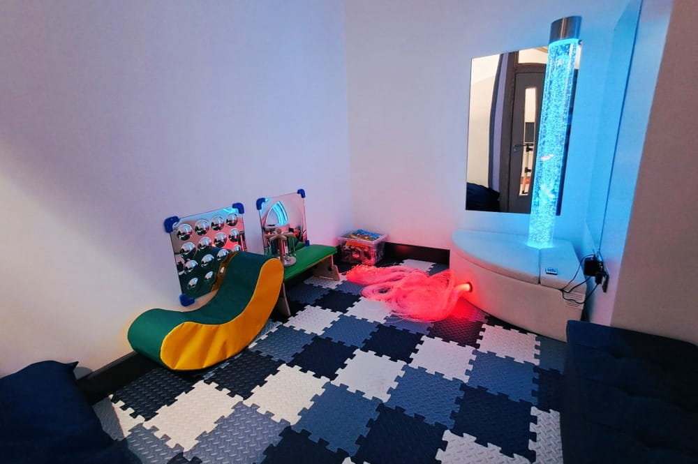 A sensory room, with square patterned floor, mirror, bubble light tower, and some toys and seating.