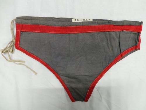 A pair of mens swimming briefs from the 1920s, made of grey material with a red embroidered outline.