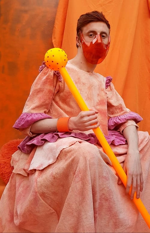 George Mohl dressed in a renaissance-style dress made of light orange corduroy and holding a sphere-topped yellow stick.He is wearing an orange plastic mask over his mouth.