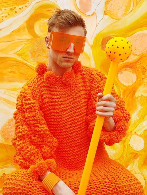 George Morl dressed in a knitted orange outfit with pompoms at the neck and wrists. He is holding a yellow sceptre and has a pair of large orange glasses on his face.