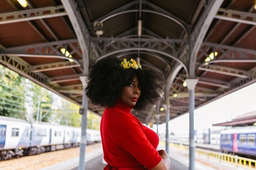 Elsa James, a Black woman in a red velvet dress and wearing a small gold crown, standing in the middle of a train station platform looking back towards the camera.