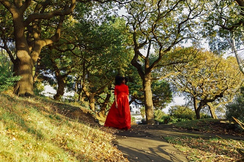 Elsa James, a Black woman in a red velvet dress and wearing a small gold crown, standing on a path through some grass, surrounded by trees.