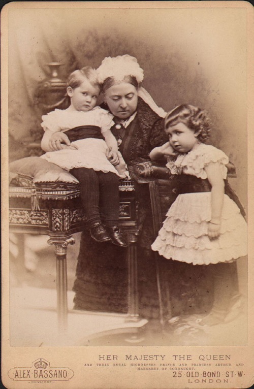 Antique postcard depicting Queen Victoria with her grandson Arthur of Connaught, wearing a frilly dress and ankle boots, and her granddaughter Maragaret of Connaught in a similar outfit.