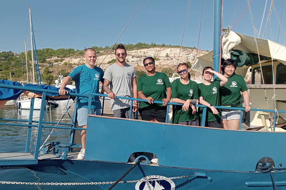 A group of six students standing on the side of a boat in Greece on a bright sunny day.