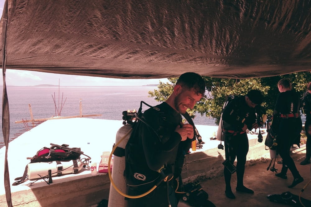 A young man dresed in diving gear adjusting an oxygen tank, two more students in diving gear visible in the background on the right.
