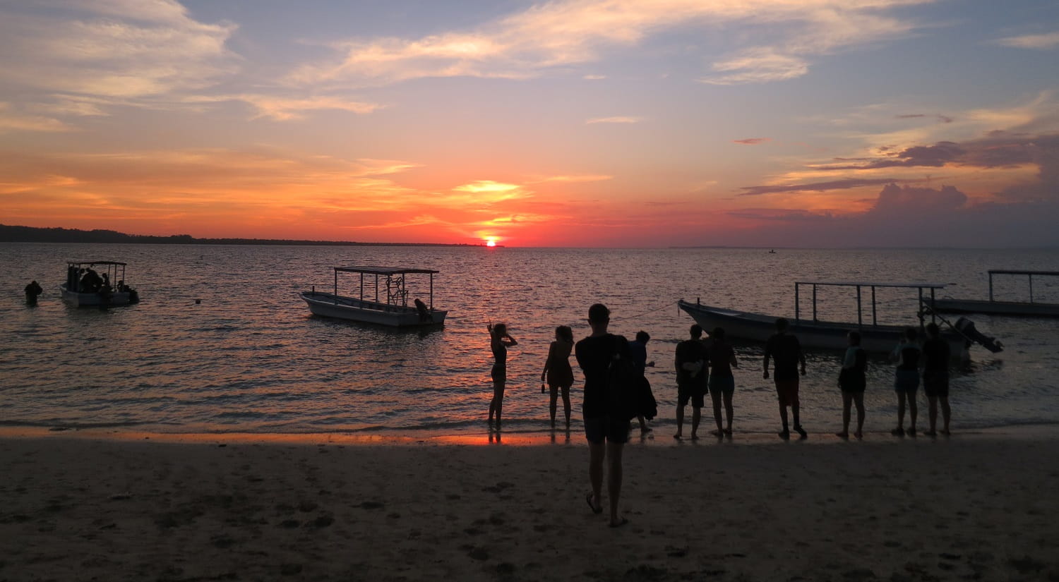 Silhouettes of students on a beach with an orange sunset in the distance across the water.