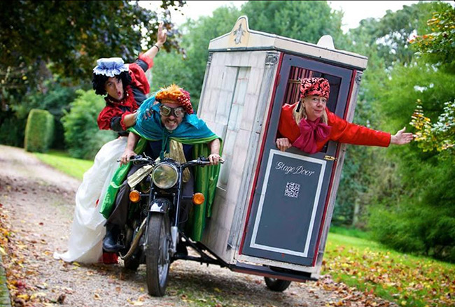 Three people all dressed in quirky, colourful outfits, with two people riding on a motorbike and one in a sidecar. Except instead of a sidecar there is a large building type box with the words 'stage door', and the person is hanging out the door with their arm outstretched.