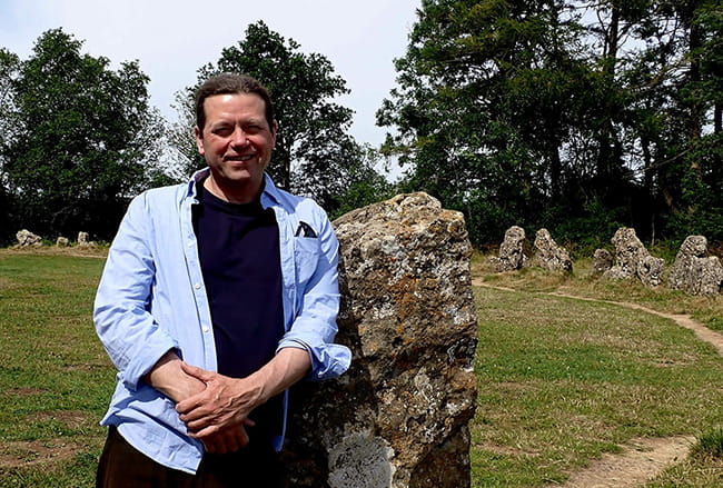 James Canton is looking to the camera and smiling, whilst leaning on a large boulder. In the background are trees, grass and more boulders that are embedded into the ground.