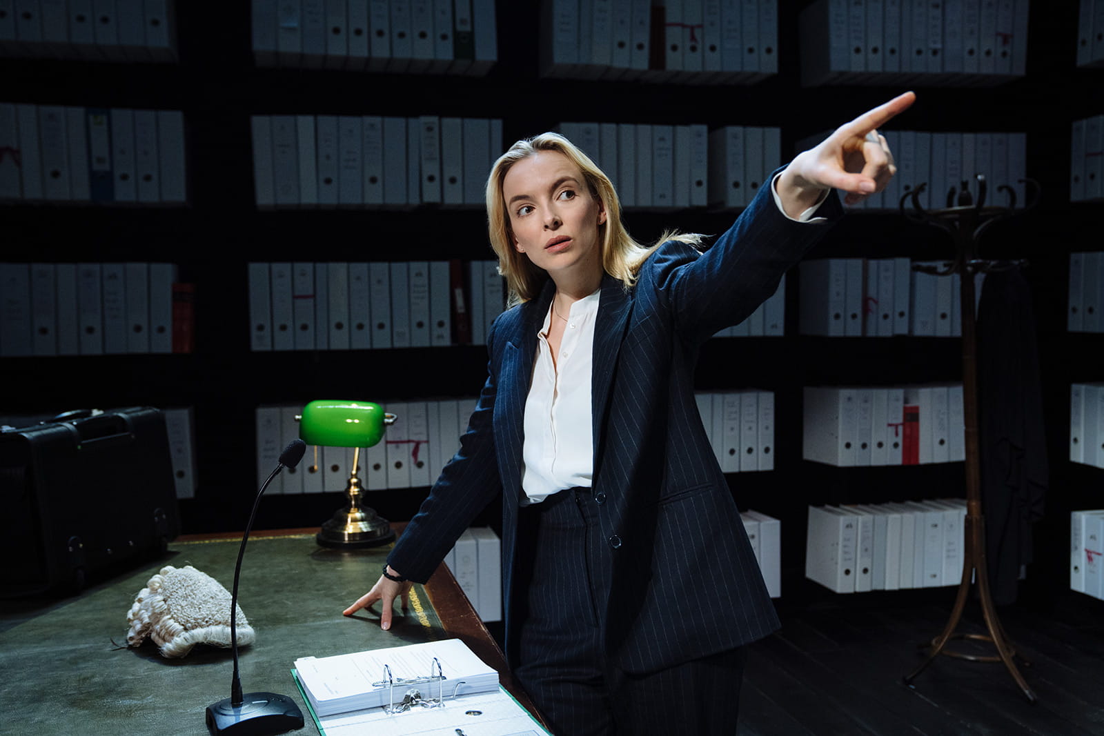 Jodie Comer as Tessa in Prima Facie. Tessa is dressed smartly in a suit, and is pointing off into the distance. Tessa's other hand is leaning against a green leather topped desk, and there are shelves of files behind her.