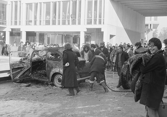 A black and white archival photograph from the University of Essex. A burnt car is stationary on Square 4, near the then-fountain, as onlookers gather around the car. 