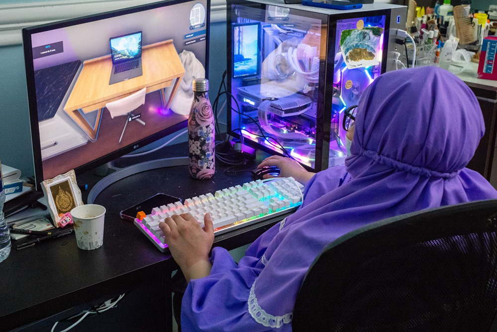 Artist Farah Al Qasimi is sat at a desk, with their back towards the camera. The focus of the photograph is instead a computer where Farah is playing a game called 'House Flipper', on the computer screen is a snapshot of the game - a computer desk and a laptop left open and unattended. 