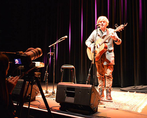 A performer on stage, singing into a mic while playing a guitar.