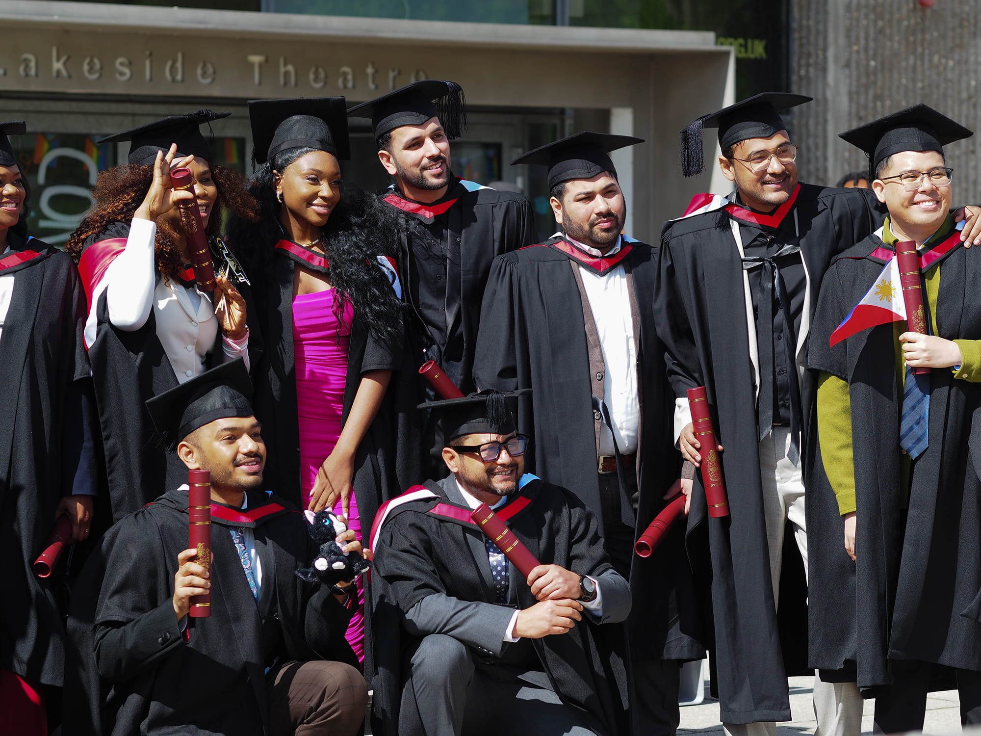 A group of graduates posing for a photo that's being taken out of shot. The smiling graduates are all dressed in black caps and gowns, holding their red diploma tubes. 